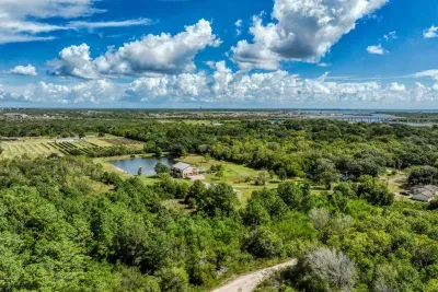 An expansive aerial view of the property and its surroundings. The home, pond, and outbuildings sit within the large, private acreage. Enjoy the mix of open fields and wooded areas, with the bay and city visible on the horizon.