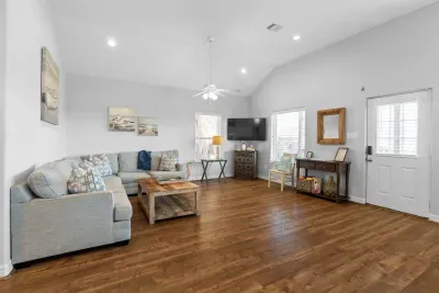 This spacious, beach-themed living room features vaulted ceilings, a large gray sectional, a rustic coffee table, dark hardwood floors, and coastal artwork, creating a bright and comfortable retreat.