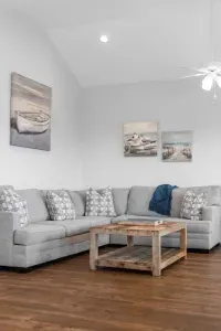 This spacious, beach-themed living room features vaulted ceilings, a large gray sectional, a rustic coffee table, dark hardwood floors, and coastal artwork, creating a bright and comfortable retreat.