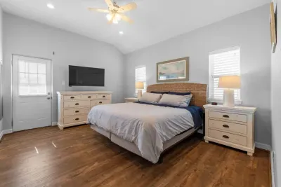 Enjoy this spacious master suite with a vaulted ceiling! A comfortable king bed, stylish rattan headboard, and calming coastal decor are complemented by a large mounted TV and ample clothing storage  in the matching white dresser.