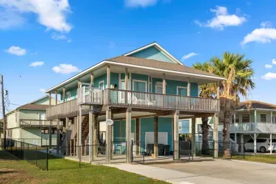 Welcome to At Last! This vibrant turquoise, two-story raised coastal home features a large Upper Deck/Balcony and a covered ground-level patio, perfect for outdoor living.
