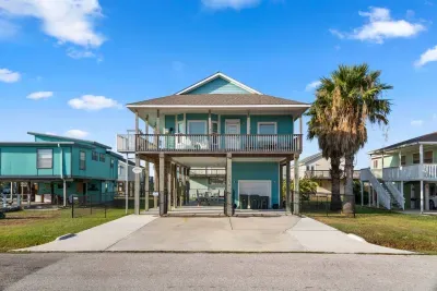 Welcome to At Last! This vibrant turquoise, two-story raised coastal home stands out with a large Upper Deck/Balcony and a covered ground-level patio, setting the stage for the perfect outdoor vacation.