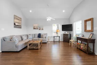 This spacious, beach-themed living room features vaulted ceilings, a large gray sectional, a rustic coffee table, dark hardwood floors, and coastal artwork, creating a bright and comfortable retreat.