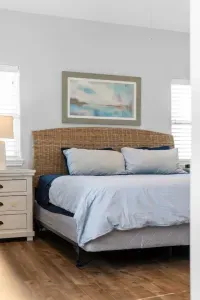 This inviting bed features a striking woven rattan headboard and calming coastal artwork. It is flanked by matching nightstands and lamps, ensuring a restful and stylish retreat.
