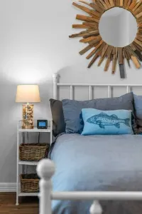 This charming room features a white metal bed with cozy blue bedding, a striking sunburst mirror, and nautical decor.