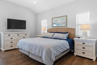 Enjoy this spacious master suite with a vaulted ceiling! A comfortable king bed, stylish rattan headboard, and calming coastal decor are complemented by a large mounted TV and ample clothing storage  in the matching white dresser.