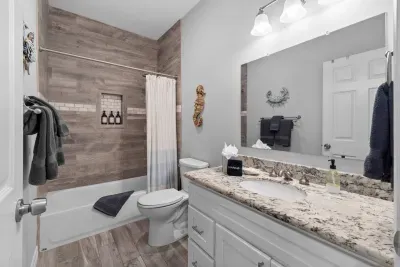 This bright, modern full bathroom features a granite-top vanity and a clean tub/shower combo. Stylish wood-look tiling, charming coastal decor, and stocked essentials and towels  ensure a refreshing guest experience.