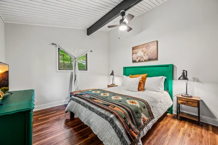 Emerald headboard against white shiplap, woven throw, smart TV on the dresser. Two windows to the woods.