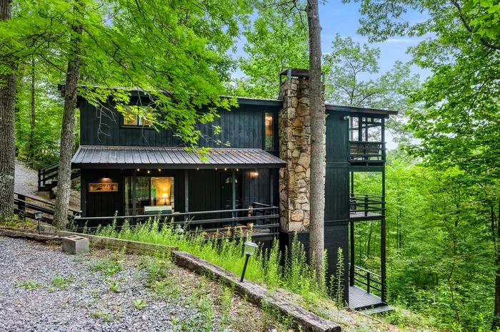 Two stories of black cedar tucked into the trees, with a stone chimney running the full height and a screened lookout on the ridge side. This is the whole cabin, from the driveway.