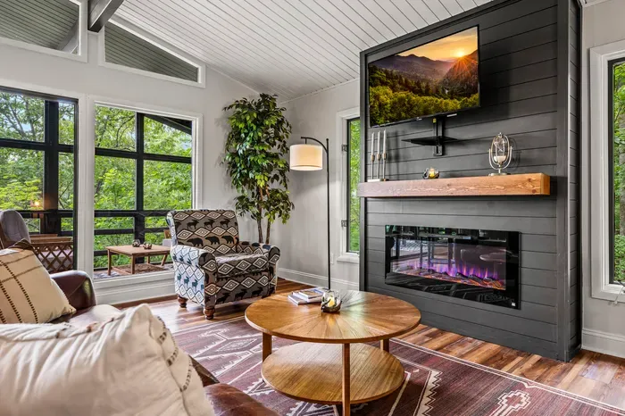 Electric fireplace under a live-edge mantel, smart TV above, and the screened porch just through the glass. Bear-and-moose armchairs pulled up close, because that's the whole point of a cabin.