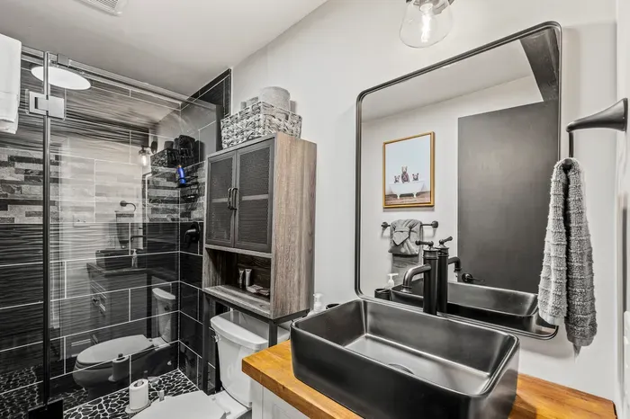 Glass walk-in shower in black subway tile with pebble floor, vessel sink on a butcher block vanity. Towels stocked.