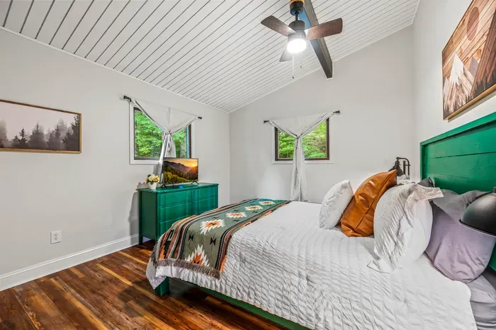 Emerald headboard and matching dresser against white shiplap, woven throw across the foot of the bed. Two windows to the woods, smart TV, and a reading lamp on the wall.