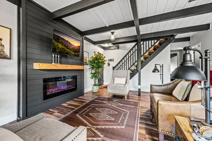 Electric fireplace under a live-edge mantel, smart TV above, open-tread stairs to the upper level. This is the room everyone ends up in.