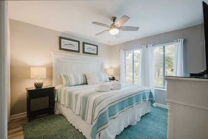 King bed dressed in coastal stripes, ceiling fan overhead, and fresh towels set out. Windows facing the lagoon keep it bright without feeling exposed.