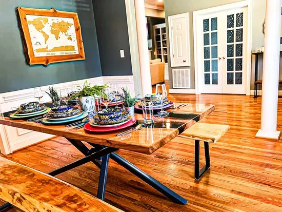 A vibrant dining room features a polished live-edge wood table set with colorful dishware and sparkling glassware. Modern benches flank the table, while potted greenery adds charm. Warm wood floors and French doors complete the inviting space.