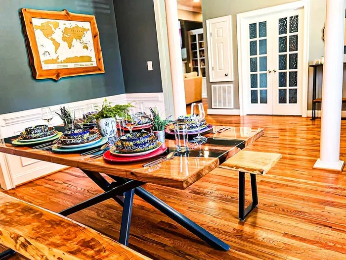 A vibrant dining room features a polished live-edge wood table set with colorful dishware and sparkling glassware. Modern benches flank the table, while potted greenery adds charm. Warm wood floors and French doors complete the inviting space.
