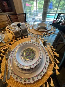 Bright and inviting breakfast place settings feature vibrant, patterned dishes layered on colorful chargers. Clear glasses, fresh greenery, and polished silverware sit atop a sleek glass table, creating a cheerful and elegant morning ambiance.