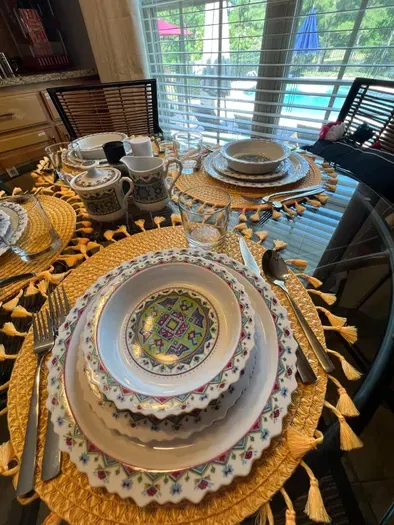 Bright and inviting breakfast place settings feature vibrant, patterned dishes layered on colorful chargers. Clear glasses, fresh greenery, and polished silverware sit atop a sleek glass table, creating a cheerful and elegant morning ambiance.