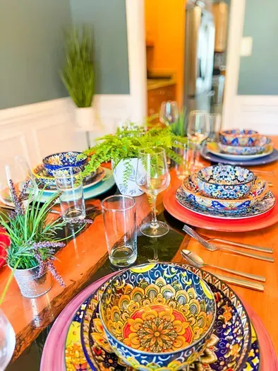 A close-up of a beautifully set table showcases vibrant, painted ceramic dishes with fractal patterns in rich blues, reds, and golds. Crystal-clear glasses, polished silverware, and lush greenery add elegance and charm to the colorful tablescape.
