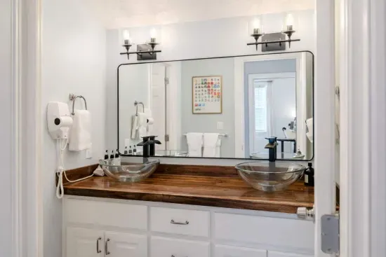 A striking black walnut live-edge slab vanity with rich, natural grain and 2 vessel sinks. Paired with a sleek mirror and warm lighting; a touch of organic elegance to the modern, spa-inspired space. Attached is a separate room with toliet & shower.