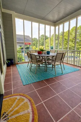 A large back porch has a sturdy, large dining table surrounded by chairs, set for outdoor gatherings. Warm natural light and greenery create a peaceful, open-air dining space perfect for  morning meals or evening relaxation. 