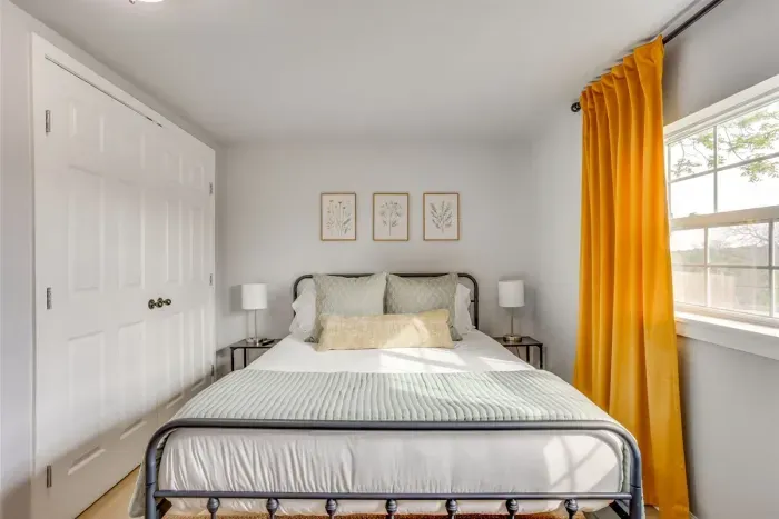 Bright guest bedroom with queen bed, iron bed frame, bold gold curtains, and large windows — warm, stylish, and comfortable.