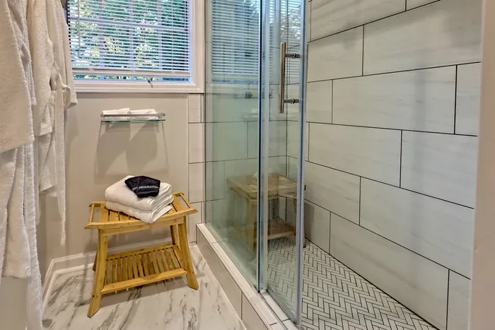 Unwind in this serene bathroom at Helen Waldhaus, featuring a walk-in shower with a spa-like bench, elegant tilework, and plush towels. The large window lets in natural light, creating a relaxing atmosphere for your stay.
