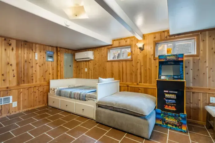 Cozy and inviting basement retreat with warm wood paneling and modern comforts! This space features a comfortable daybed that pulls out from a twin to a king, a plush oversized ottoman, and a classic Marvel arcade game for some nostalgic fun.