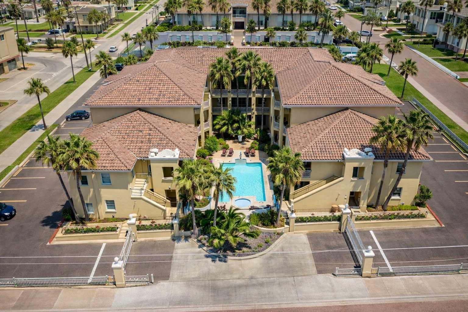Mediterranean-style retreat featuring twin stucco buildings with red-tiled roofs, a sparkling turquoise pool framed by palms, and gated entry for privacy. Ideal for families or groups seeking resort-style comfort near coastal attractions and vibrant