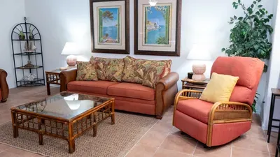 Bright, welcoming living area featuring a plush sofa, tropical‑leaf pillows, warm lighting, and a comfy recliner. Natural textures, soft tones, and thoughtful décor create a relaxing coastal retreat perfect for unwinding after a day out.