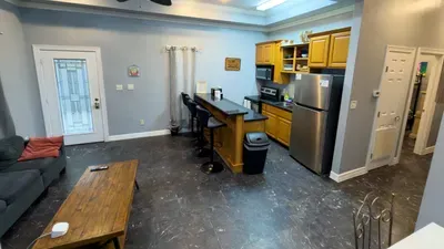 Open Kitchen-Living w/ Bar Seating
Bright, flowing space blends kitchen & lounge! Golden cabinets, SS fridge, granite bar for 2+ stools, comfy gray sofa nearby. Ideal for casual meals & hangs—fully equipped for fun!