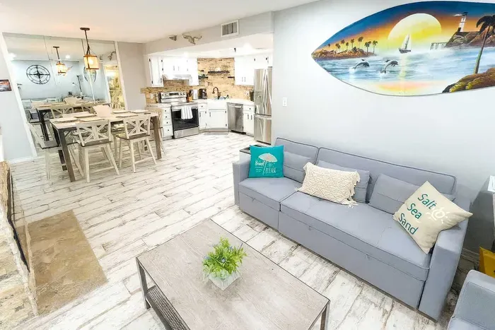 Unwind in a stylish, beach-inspired living space with plush seating, coastal decor, and an open layout. The nearby dining area and fully equipped kitchen make entertaining easy, while mirrored accents enhance the breezy, light-filled ambiance.