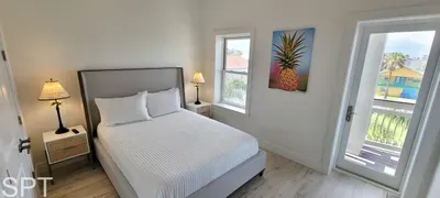 Airy coastal bedroom with soft white linens, warm wood floors, and balcony access overlooking palms. A bright, calming retreat designed for slow mornings and breezy evenings.