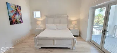 Bright coastal bedroom with crisp white décor, plush bedding, and private balcony access. Sun‑filled space with serene artwork, warm wood floors, and relaxing tropical views—perfect for unwinding after a beach day.