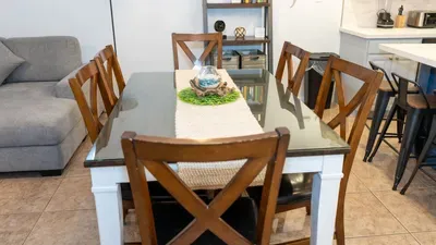 Bright Open Kitchen & Dining Retreat.
Spacious white shaker cabinets, stainless appliances, navy island flow into wood farmhouse table seating 6 under industrial pendant lights. Cozy gray sofa, plants, lamps—ideal for family meals.
