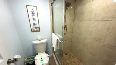 Coastal Bliss Spa Bathroom w/ Walk-In Shower. Unwind in this serene coastal-inspired bathroom featuring a spacious walk-in shower with rainfall head, crisp white tiles, fluffy towels, and nautical lighthouse art. Perfect for refreshing stays!