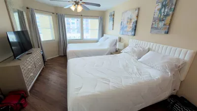 Bright and inviting double‑bedroom retreat featuring crisp white linens, ample natural light, a sleek dresser with smart TV, and calming neutral tones—perfect for families or groups seeking comfort, space, and a relaxing stay.
