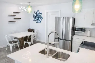 Bright Coastal Kitchen + Dining | Stylish Decor, Stainless Appliances, Blue Sun Accent, Floating Shelves, Modern Lighting, Family-Friendly Layout, Walk to Beach, Ideal for Couples or Small Groups