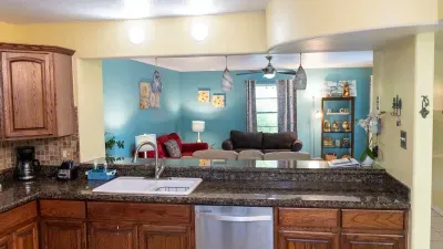 This kitchen features rich wood cabinetry, granite countertops, and modern appliances, including a dishwasher. A large pass-through opens to the cozy living room, creating a bright, connected space perfect for cooking and entertaining with ease.