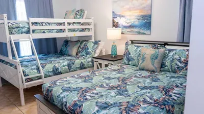 Bright coastal bunk room with a full‑over‑twin layout, tropical bedding, and soft blue accents. A cozy, well‑designed space offering comfort and convenience for families and groups enjoying a relaxed seaside getaway.