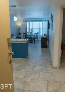 Welcoming entryway opening into a bright living and dining space with tiled floors, a stylish blue kitchen island, warm lighting, and natural light from large windows—creating an inviting, open layout perfect for relaxed vacation living.