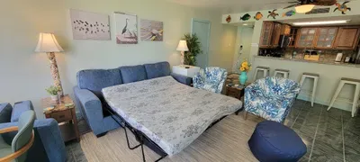 A cozy, guest‑ready living area featuring a fold‑out sofa bed, floral accent chairs, a woven rug, and a bright kitchen with bar seating, creating a comfortable, functional space perfect for relaxing, gathering, and enjoying easy vacation living.