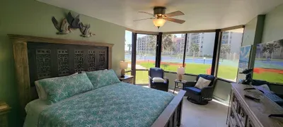 A bright, inviting bedroom featuring a carved wood headboard, green patterned bedding, cozy seating by expansive corner windows, and views of the courts and skyline, creating a relaxing, sun‑filled retreat perfect for unwinding in comfort.
