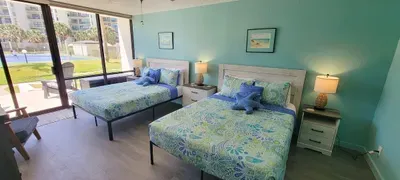 A bright, coastal‑themed double‑bed retreat featuring ocean‑inspired bedding, soft turquoise walls, charming décor & sliding doors that open to a sunny patio with pool views, creating a relaxing, family‑friendly space.