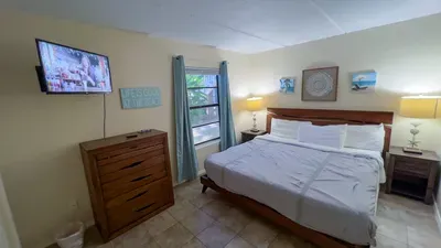Coastal Serenity Master Bedroom
comfy bed w/ crisp white linens anchors this airy retreat featuring wall-mounted TV, wood dresser, bedside lamps & beach wall art. Blue drapes frame window views "Beach" sign enhances the relaxed, seaside escape vibes.