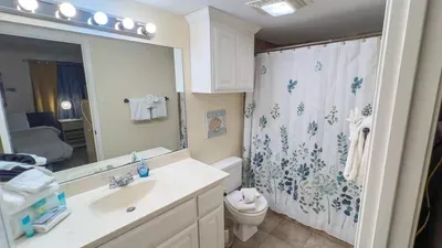 Floral Coastal Guest Bathroom
Clean white vanity, toilet, and walk-in shower with blue floral curtain greet you. Bright vanity lights, fluffy towels, storage cabinets, and beachy wall art provide a fresh, spa-like retreat for guests.