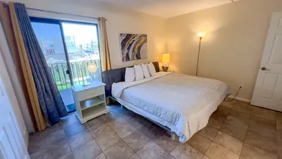 Balcony View Bedroom
Serene bed with plush white duvet sits in soft light from balcony sliders with blue-yellow drapes. Nightstand lamp, abstract art, and balcony overlook offer tranquil coastal mornings and serene evenings.