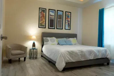 Relax in this bright, modern bedroom featuring a plush upholstered bed, layered pillows, warm wood flooring, and soft natural light. A cozy reading nook, vibrant artwork, and calming blue accents create a welcoming, restful retreat.