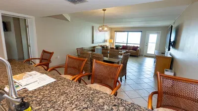 Bright open‑concept living space featuring a granite breakfast bar, cozy dining setup and inviting lounge with natural light streaming through large windows. Perfect for gathering, cooking and relaxing in a warm, modern setting.