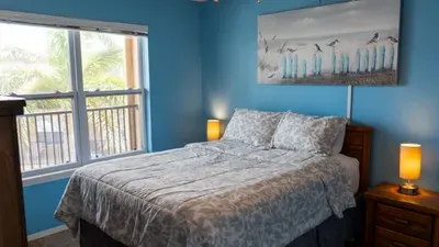 Rest comfortably in this serene bedroom, featuring a plush comfy bed, cozy bedding, and tranquil blue walls. Large windows let in natural light and palm views, making it an inviting retreat after a day at the beach.